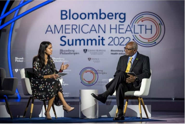 On a stage sitting in chairs, Environmental Justice Coalition Founder Rhea Goswami interviews Professor Robert Bullard. The wall behind then has text: Bloomberg American Health Summit 2022. 