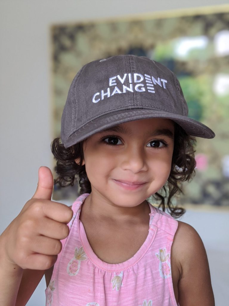 A young girl wearing a gray hat with the words Evident Change gives a thumbs up. 