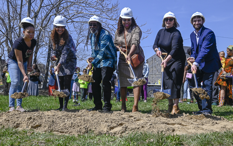 A group of five people wearing white hard hats each lift a silver shovel with dirt on a plot of green grass.