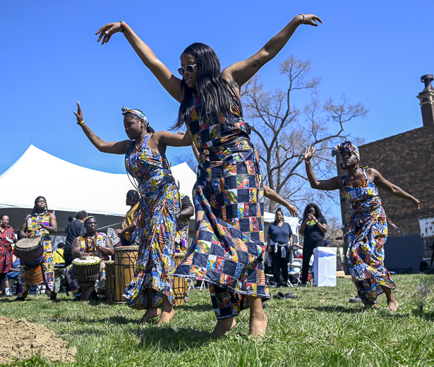 Three women wearing patterned blue, red, yellow and green sleeveless dresses perform a dance on green grass in front of a white tent. 