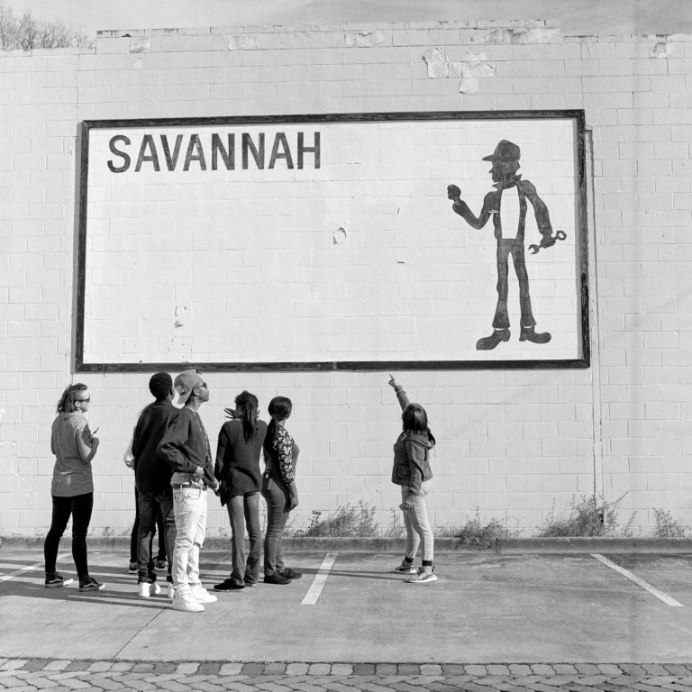 A black and white image shows a group of youth looking up and pointing at a large mural. The mural is a drawing of a rectangle with the word Savannah written in the top left-hand corner, a large blank space underneath the word, and a man holding a wrench in the right-hand corner. 