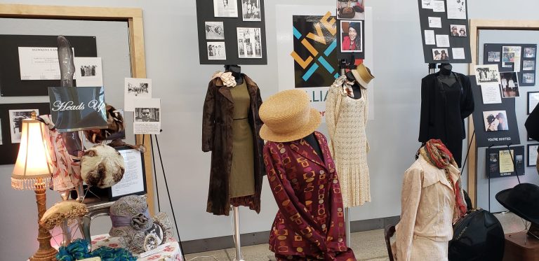 Items in exhibition include fashionable dresses and women's hats on manneqiuns. Photos are on walls but are too small to make out.