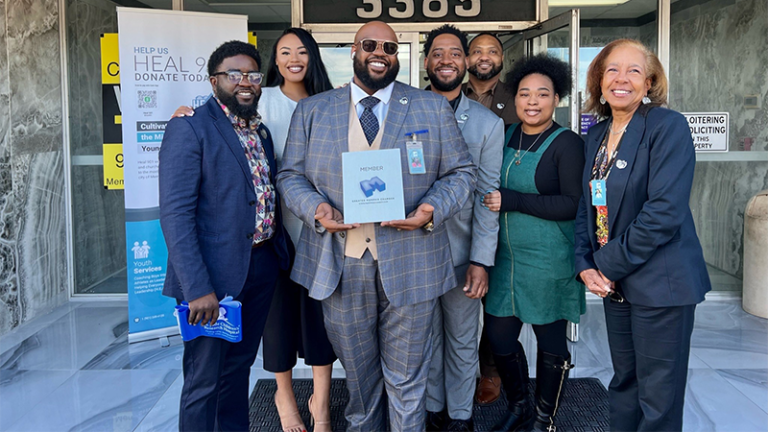A group of seven people are see posing in front of a building with a man in a suit holidng a membership certificate to the Greater Memphis Chamber.
