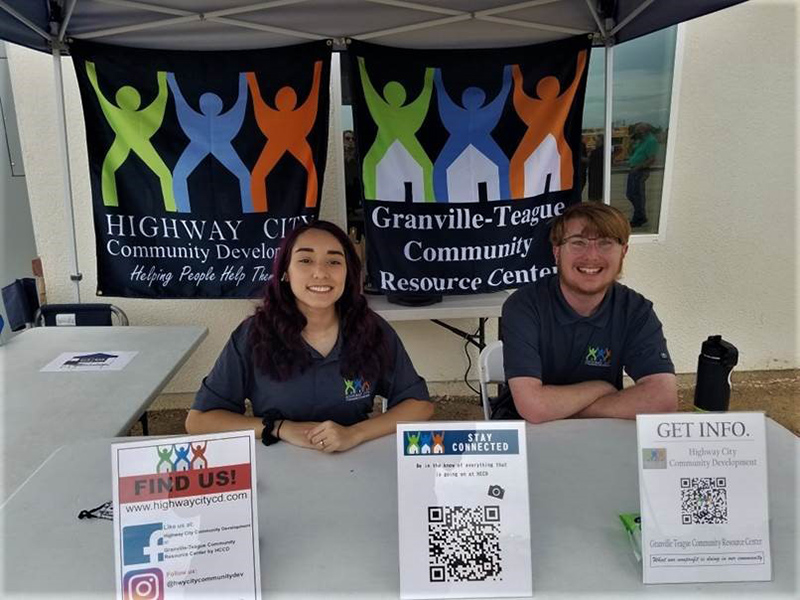 Two people sit at a desk with signs behind them that read: ‘Highway City Community Development Helping People Help Themselves’ and ‘Granville-Teague Community Resource Center.’ On the desk includes material about the organizations on how to ‘Find Us,’ ‘Stay Connected’ and ‘Get Info.’