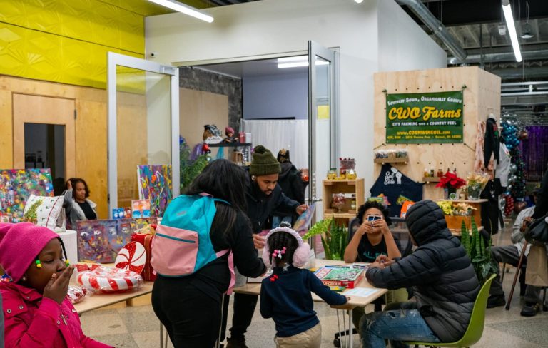 Large room with small child in winter jacket in foreground, children and a teen or young adult at a table playing board games in the middle ground and vendors in the background