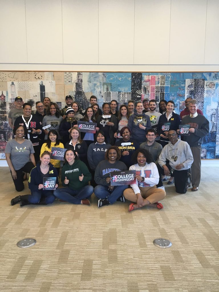 A group photo of Kresge staff celebrating College Signing Day by wearing college gear and holding Better Make Room #CollegeSigningDay .