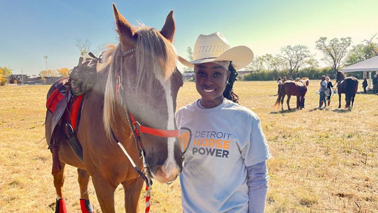 A woman in a cowboy hat and Detroit Horse Power T-shirt stands next to and holds the reins of a horse.