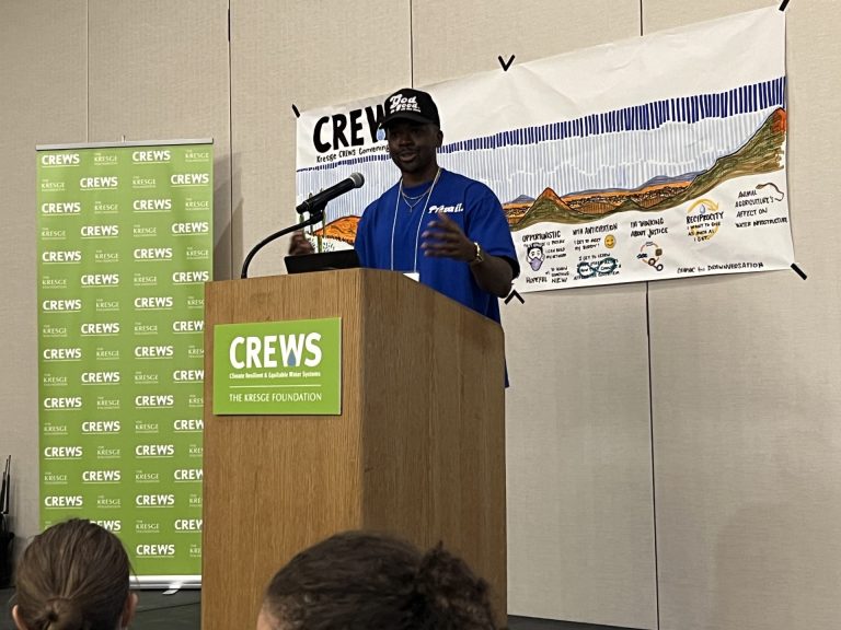 Hip-Hop artist and filmmaker Benny Starr in a black God Is Good hat and blue shirt with the word Prevail on it, stand behind a lectern with a CREWS Climate Resilient and Equitable Water Systems sign on it and a CREWS illustration behind him.