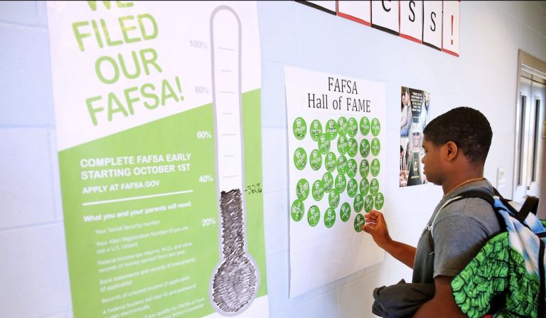 A college student stands in front of a wall with poster that say: "We filed our FAFS!" and FAFSA Hall of Fame."