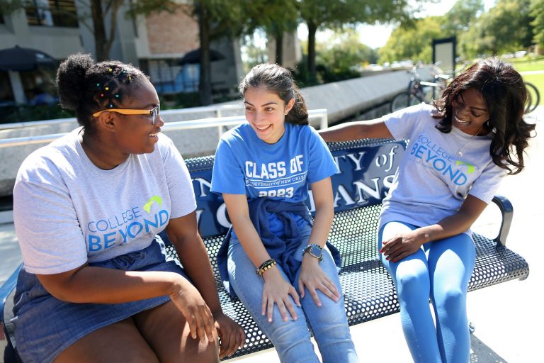 Three students sit on a bench and talk to each other. Two wear t-shirts that read 'College Beyond.'