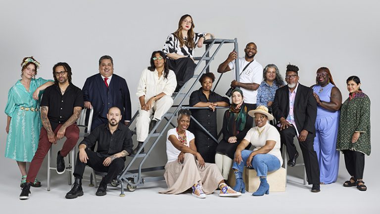 A large group photo of 15 artists with several standing and ithers sitting on chairs and a movable metal staircase.