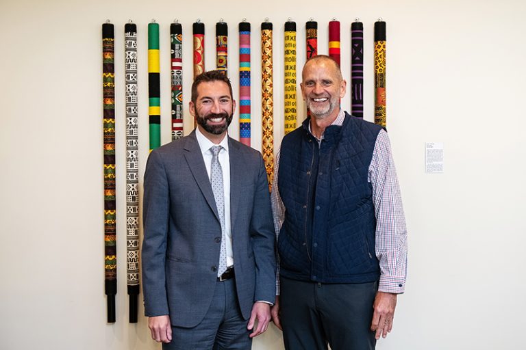 A photo of two white men standing before a mixed media work of several long and narrow multi-colored art objects.