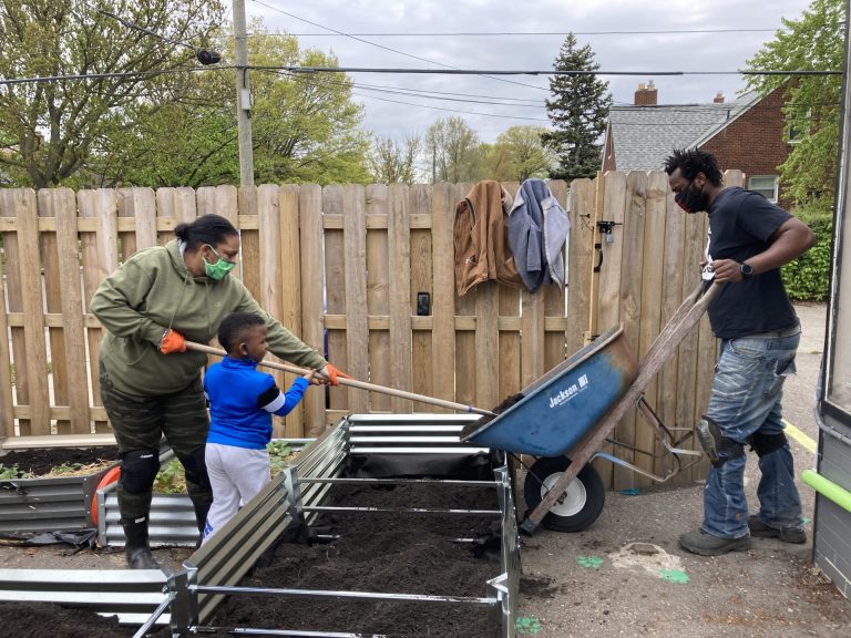 Working on outdoor raised garden beds, Woman with rake pulling material -- with a young child helping guide the rake -- from a wheel barrow being tipped by a man.