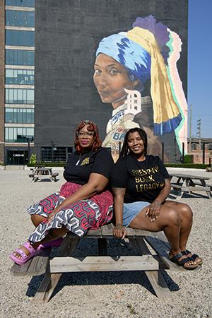 Two Black women are sitting on a picnic table in a gravel lot with a large mural in the background of a Girl with a D Earring Mural. 