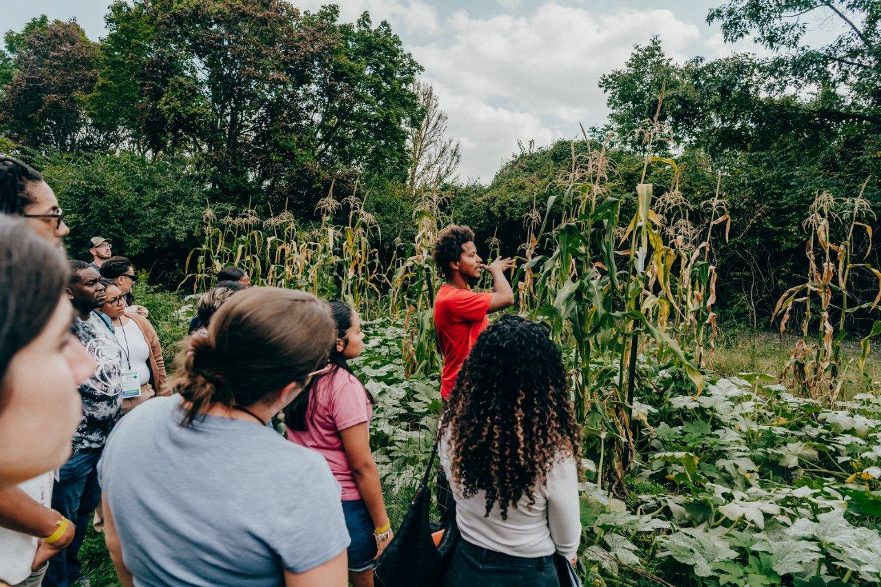 A man stands in a garden pointing to the plants in front of a group of people visiting a farm.