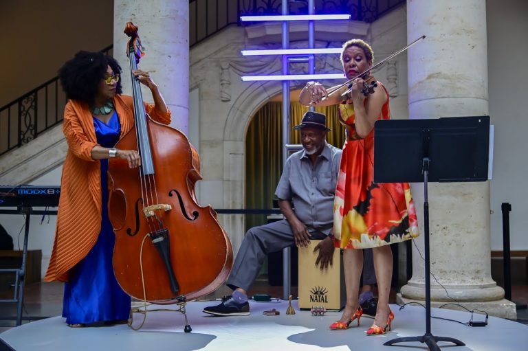 Marion Hayden in a blue dress and orange coat is playing the bass and Michelle May in a colorful dress is playing the violin, with percussionist Mahindi Masai sitting and plahing behind them. .