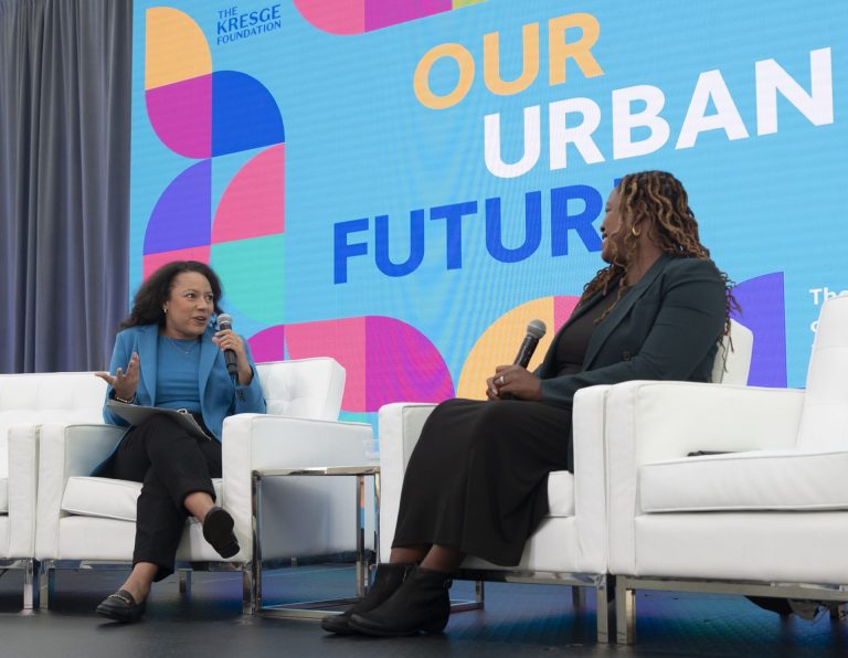 Two professional woman speak on stage at Our Urban Future. They're in white arm chairs and both are women of color.