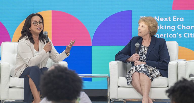 Two women sitting in white chairs on a stage are having a discussion at the Our Urban Future convening.