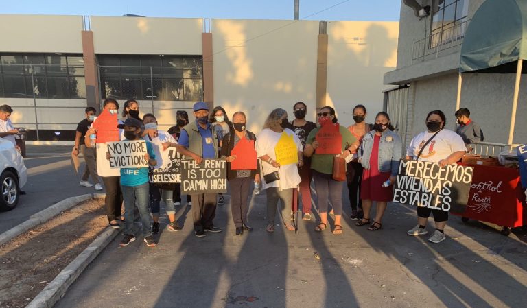 A group of 15 union members wearing face masks stand in a parking lot with signs that read "no more evictions," "the rent is too damn high" and "merecemos viviendas estables."