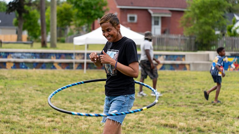 Several adults and kids play in a park, including one using a hula hoop