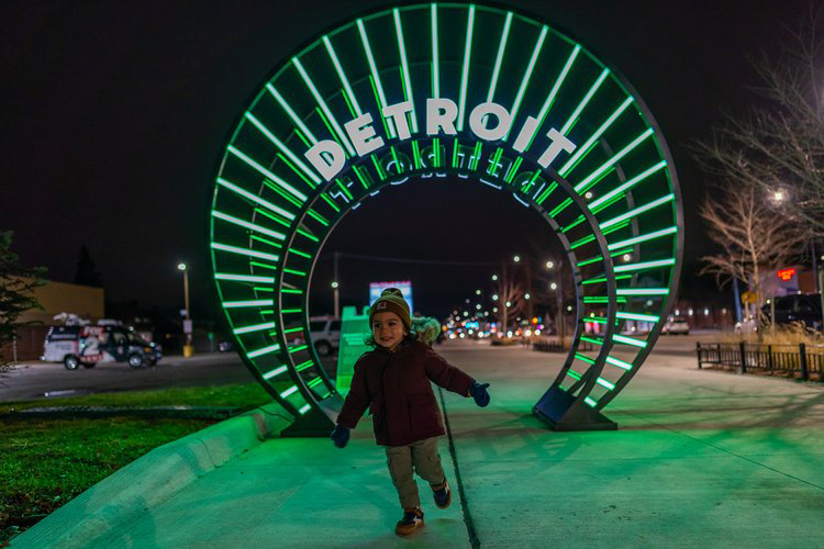 A young child in a winter jacket and hat is playing in front of a green lit-up circular sign with the word Detroit on it.