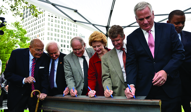 A line of seven people (government, city and philanthropic leaders) are signing a steel beam at a construction site.