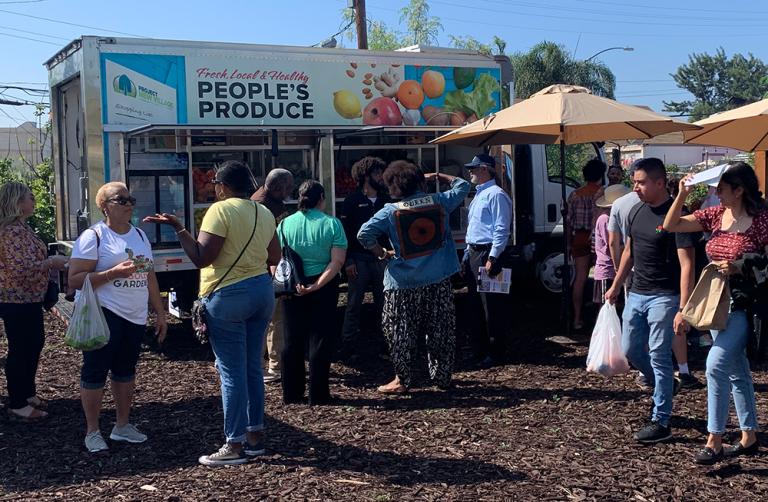 A crowd of people gather in front of a white food truck that says People's Produce on the side.