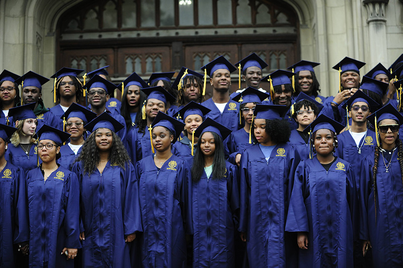 A group photo of graduating high school students in cap and gown standing in front of the school building at the School at Marygrove.