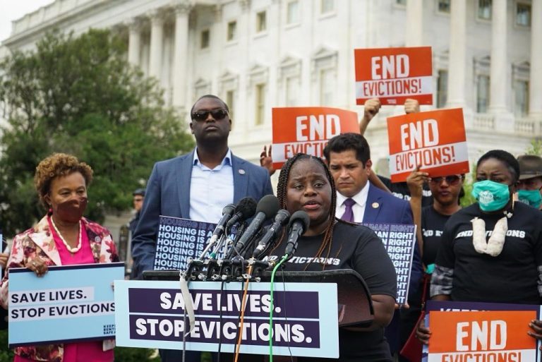A woman with long hair stands at a podium with microphones and a sign that reads "Save Lives. Stop Evictions." with a group of seven people behind her.