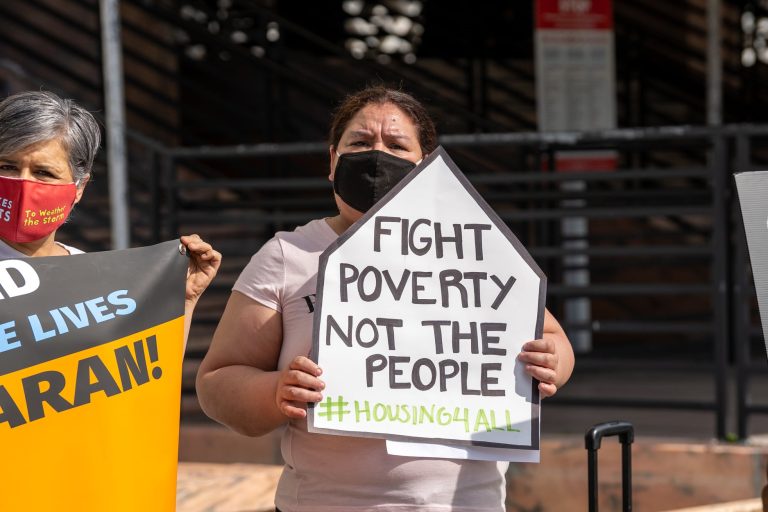 A woman wearing a black mask stands with a white sign reading "Fight Poverty, Not the People, #Housing4All."