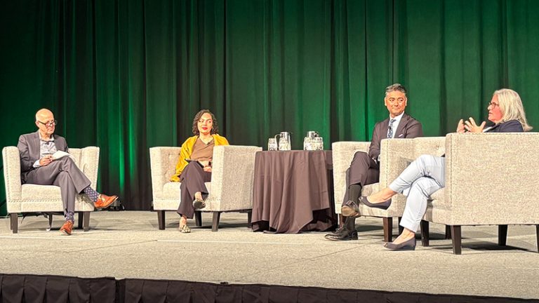 Four speakers on a panel sitting on large white chairs on a stage with a green curtain in the background