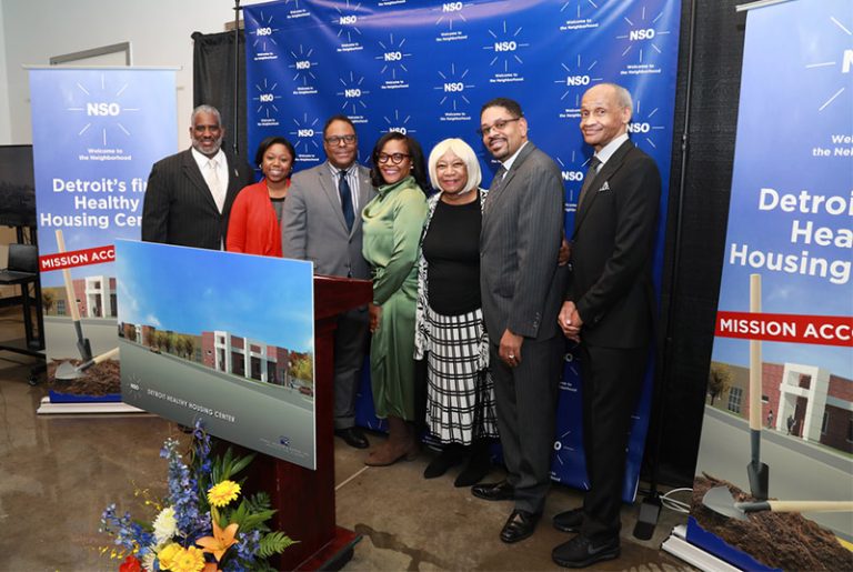 A group of 7 people are lined up at a presentation with a photo of the Detroit Healthy Housing Center in front of them and a NSO blue screen behind them.