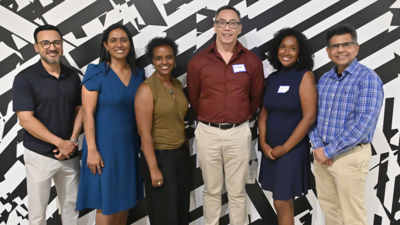 Six people are standing in front of a black and white striped wall.