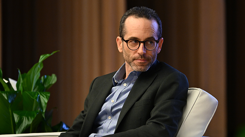 Brett Theodos, a senior fellow and director of the Community Economic Development Hub at the Urban Institute, sits in a chair on a stage next to a plant.