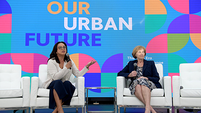 Two women with microphones in hand sit in white chairs on a stage with an Our Urban Future graphic on a screen behind them. 