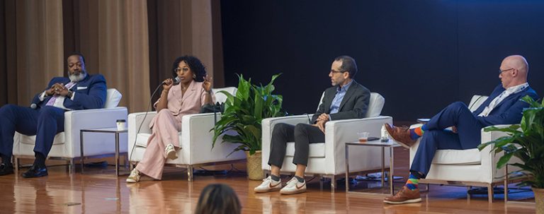 A panel of four speakers sitting on a stage in white chairs with plants between the chairs.