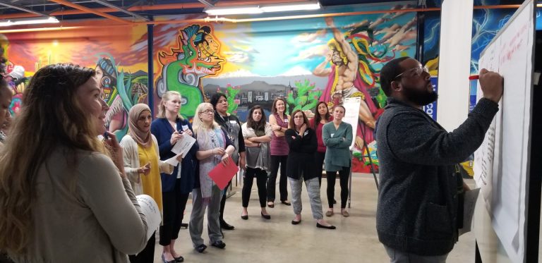 Bright Mexican-themed murals are in the background while a dozen women in an open meeting area watch a man write on a dry erase board,