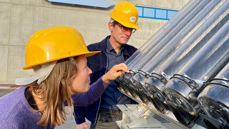 A man and a woman in yellow hard hats inspect solar equipment at energy plant.