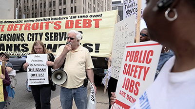 A group of people at a protest with signs saying: "Jobs, Pensions, City Services, The Banks Owe Us."