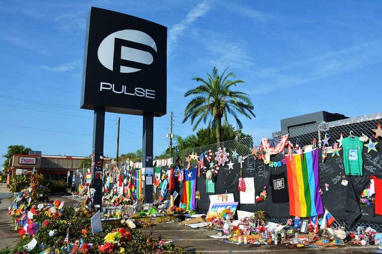 A scene of the Pulse nightclub in Orlando, Florida in 2016 after the deadliest attack on the LGBTQ+ community on our nation’s history. A large number of memorials to the victims of the shooting are left on the fence and ground outside the club, including flowers, signs, photos, cards, Pride and American flags.