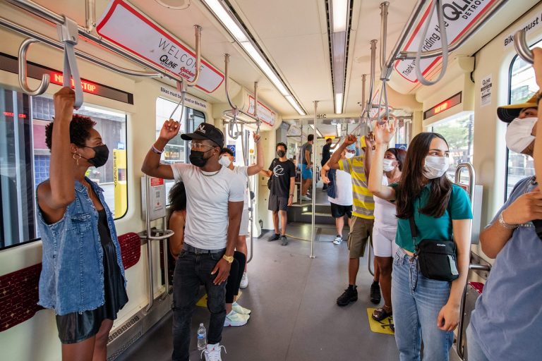 A diverse group of riders inside one of Detroit’s QLine streetcars. All of them are masked and standing, holding on to straps and rails. 
