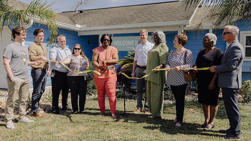 A group of nine people are standing in a line on the lawn in front of a blue house. The group is holding a yellow ribbon as the person in the middle cuts the ribbon.