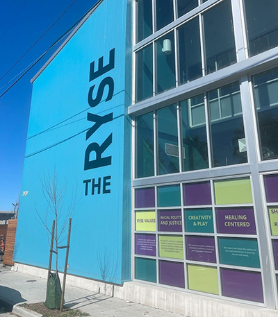 A large building with a blue wall and the words the RYSE and large windows with sayings like Racial Equity and Justice, Creativity & Play. 