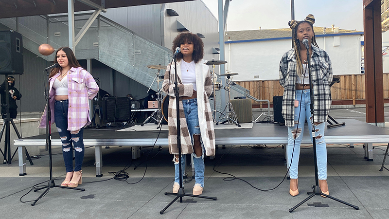 Three young people are standing on a stage in front of microphones singing.