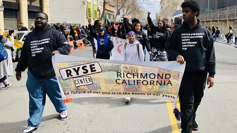 Two young people of color in black sweatshirts that say “Justice and Equity, Youth Leadership, Safety, Partnership, Creativity, Fun” carry a banner that says “RYSE Center, Richmond, California, The future of our city is in the hands of our youth,” as they lead a group of other youth down a street in a parade with onlookers on the side.