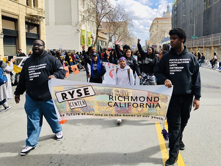 Two young people of color in black sweatshirts that say “Justice and Equity, Youth Leadership, Safety, Partnership, Creativity, Fun” carry a banner that says “RYSE Center, Richmond, California, The future of our city is in the hands of our youth,” as they lead a group of other youth down a street in a parade with onlookers on the side.