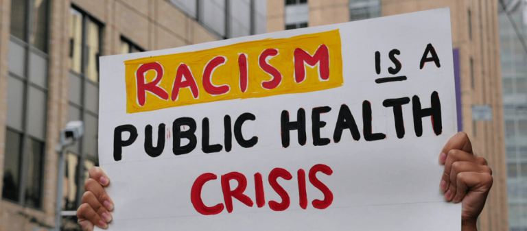 Two hands hold up a white sign reading "Racism is a Public Health Crisis" in red letters