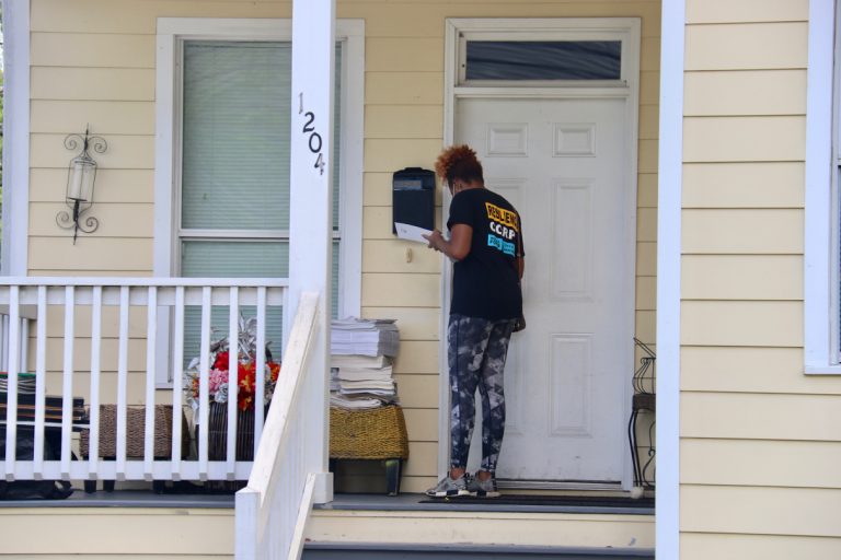A woman wearing a black t-shirt with the words Resilience Corps on it knocks on the front door of a house.