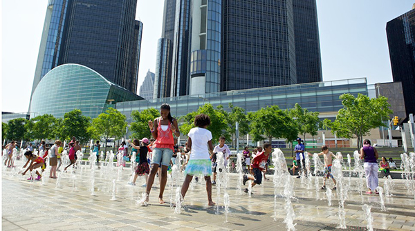 A group of young children play in a plaza with fountainsof water sprouting up in front of the glass towers of the Renaissance Center in Detroit.
