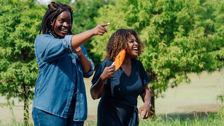 Two women, one smiling and pointing and the other laughing and holding an orange fan, are having fun at the Sankofa Wetland Park and Nature Trail.